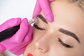 Permanent make-up for eyebrows of beautiful blonde woman in beauty salon. Closeup beautician doing eyebrows tattooing.