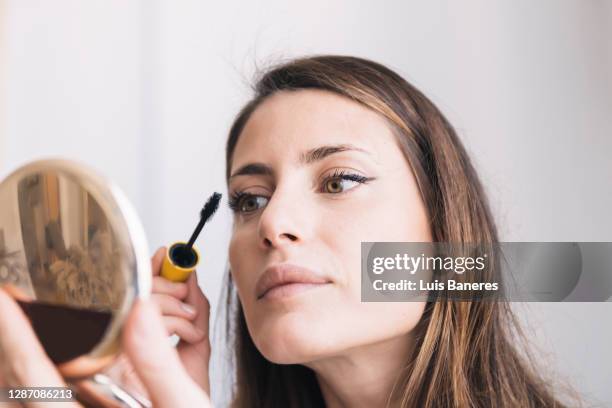 young female with compact mirror in hand applying eyeliner during daily beauty procedure at home - wimper stockfoto's en -beelden