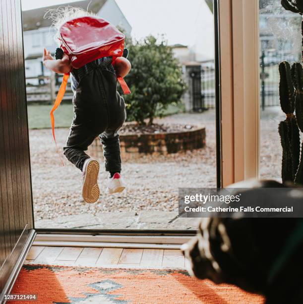 happy little girl leaps out the sunny front door, while wearing a red backpack. her dog looks on as she leaves. - eerste-schooldag stockfoto's en -beelden