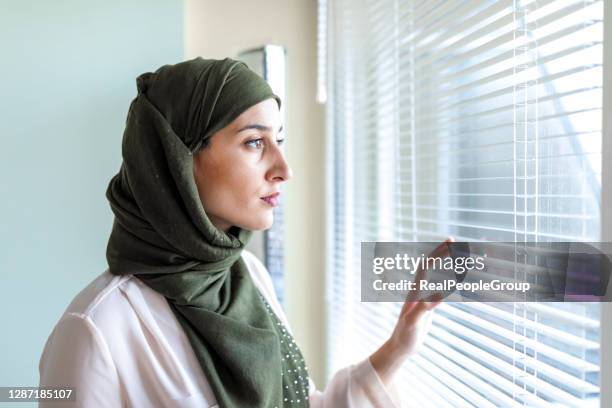 frau mit hijab steht neben fenster. besorgte muslimische junge frau, die durch ein fenster schaut - arabische frau kopftuch stock-fotos und bilder