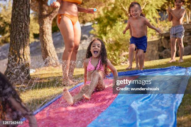 children sliding on water slide with mother on garden - backyard water slide stock pictures, royalty-free photos & images