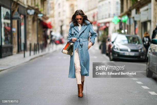 Therese Hellström wears sunglasses, a blue long winter wool trench coat with fluffy faux fur collar, white pants, a fluffy red and green khaki bag,...