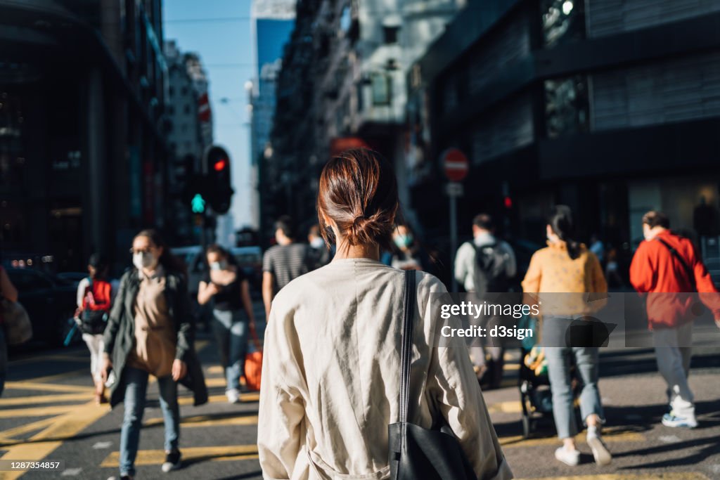 Rear view of young Asian woman commuting to work in city in the fresh morning, crossing street amidst crowd of pedestrians. Daily life and routine of a businesswoman