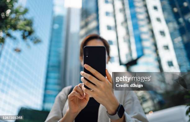 low angle view of confident and successful young asian businesswoman using smartphone in financial district, against corporate skyscrapers in the city. business on the go - viso nascosto foto e immagini stock