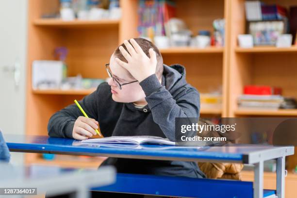 intellectually disabled boy sitting at the classroom desk and writing - special education stock pictures, royalty-free photos & images