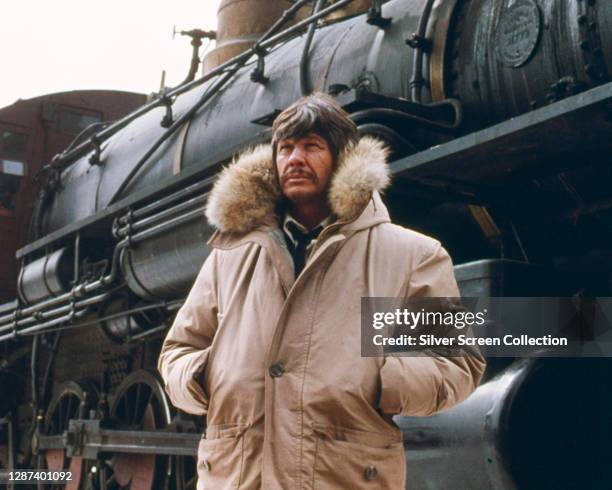 American actor Charles Bronson wearing a parka over his costume on the set of the 1975 film 'Breakheart Pass'. He stars as undercover agent John...