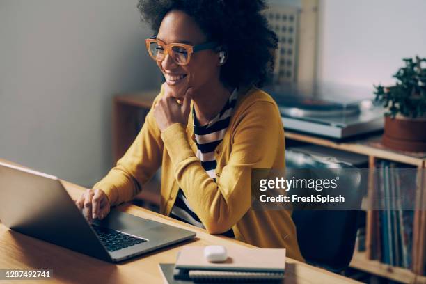 smiling african american woman wearing glasses and wireless earphones makes a video call on her laptop computer at her home office - laptop imagens e fotografias de stock
