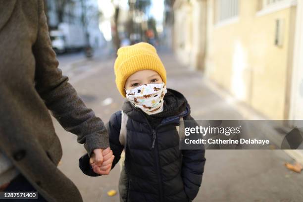 a little girl wearing a protective face mask, holding the hand of her mother in the street before to go to school - winter illness stock pictures, royalty-free photos & images