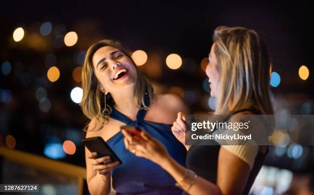 stilvolle freunde stehen nachts auf einem balkon und lachen über eine nachricht - woman in evening dress stock-fotos und bilder