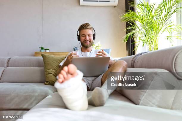 fröhlicher junger mann mit gebrochenem bein bei videoanruf - broken leg stock-fotos und bilder