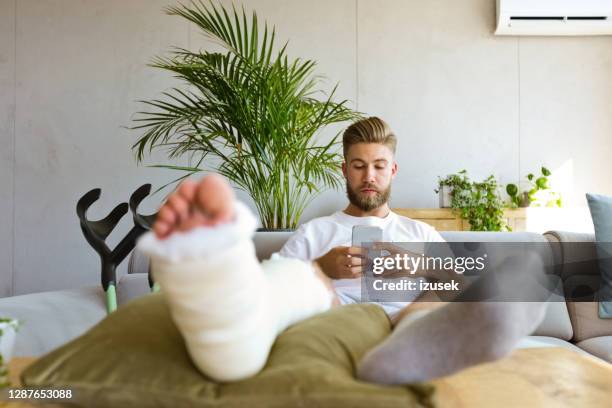 junger mann mit gebrochenem bein mit smartphone - orthopedic cast stock-fotos und bilder