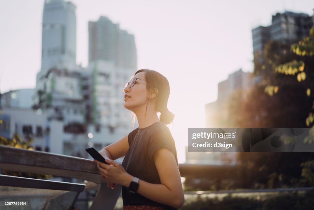 Confidence young Asian businesswoman with smartphone taking a break in urban balcony of a office building in financial district in the city, looking away while enjoying some fresh air in the middle of a busy work day