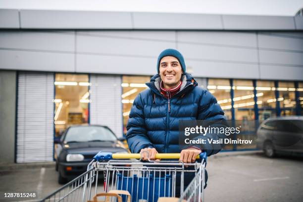 glimlachende jonge mens met een boodschappenwagentje voor een grote winkel. consumentisme / shopping concept. speciale gelegenheid / vakantie winkelen concept. - shopping trolleys stockfoto's en -beelden