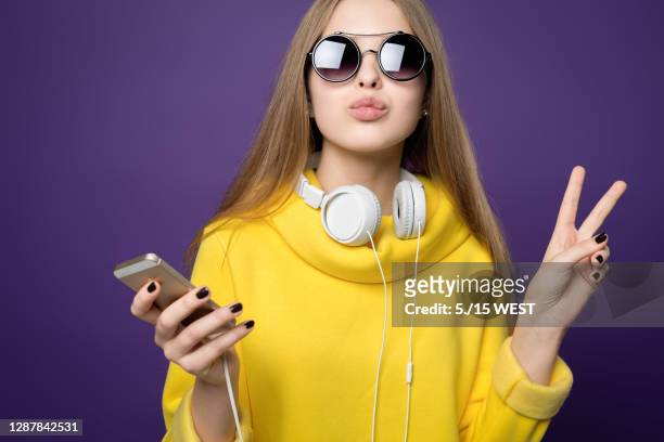 kissing peace. young beautiful girl in yellow sweatshirt and glasses. holds the phone in hand and headphones - fashionable glasses stock pictures, royalty-free photos & images