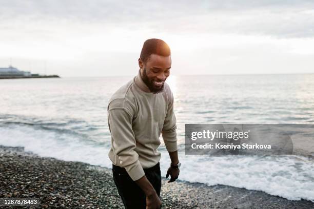 dark-skinned young man with black hair, beard and mustache walks along the beach along the sea and smiles and looks down - young male model stockfoto's en -beelden