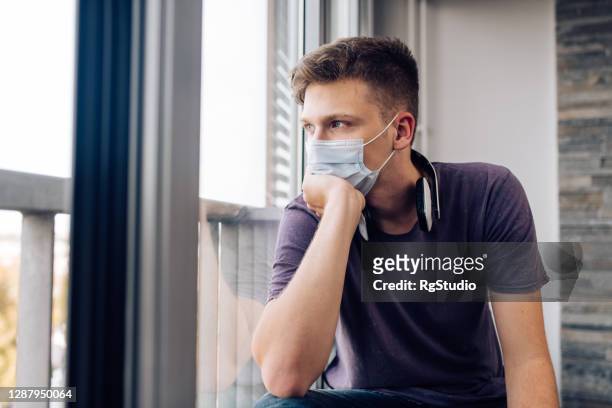 young man with a face mask sitting in a home quarantine and looking through the window - looking through window covid stock pictures, royalty-free photos & images
