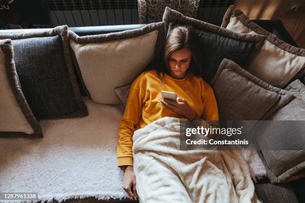 young woman lying on sofa and using cell phone at home - lying down stock pictures, royalty-free photos & images