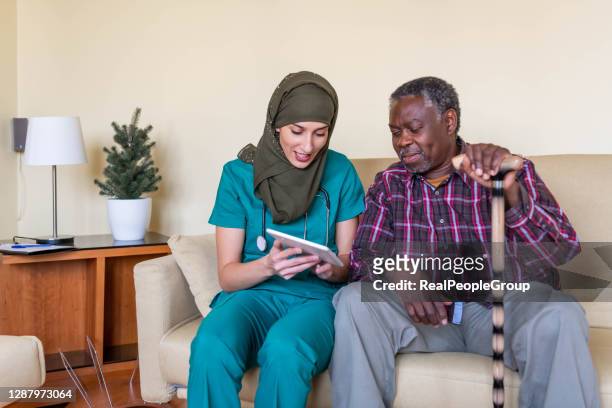 lächelnde muslimische krankenschwester hält digitale tablette von pensionierten männlichen patienten. - nurse with old male patient home care stock-fotos und bilder