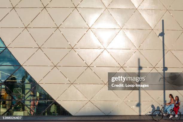 Two young women on a bicycle ride past the geometric facade of The Blob building in Eindhoven at dusk.