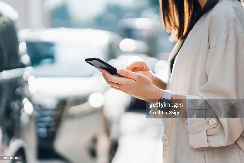 Cropped shot of chic young Asian woman using mobile app device on smartphone to order a taxi pick up service by the urban road in downtown city street. Speedy and trustworthy service. Carsharing and business on the go themes