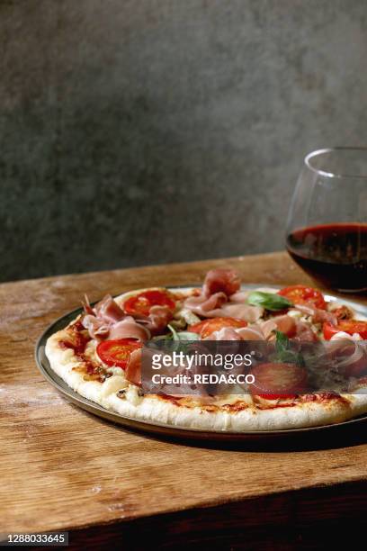 Fresh baking homemade pizza napolitana with prosciutto ham. Cheese. Tomatoes. Basil on plate. Glass of red wine over wooden table background. Home...