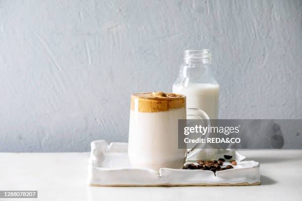 Dalgona frothy coffee trend korean drink milk latte with coffee foam in glass mug on ceramic tray with ingredients on white marble table.