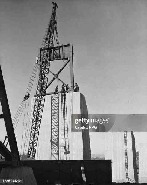 The figures of bridge workers on a concrete pier guide a steel section, raised by a crane, into place on a pier of the Pit River Bridge over which...