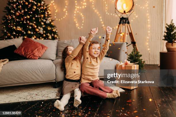 cheerful happy kids tween having fun fooling around and laughing while sitting on the floor near the decorated christmas tree sofa playing with confetti in the decorated room at home - christmas atmosphere stock pictures, royalty-free photos & images