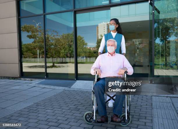 nurse pushing a senior man on a wheelchair leaving the hospital and wearing facemasks - leaving hospital stock pictures, royalty-free photos & images