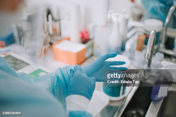asian chinese female dentist wearing protective workwear ppe in front of mirror in dentist office getting ready surgery - surgical glove stock pictures, royalty-free photos & images