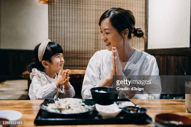 young asian mother and lovely little daughter looking face to face and smiling to each other, putting hands together in prayer, saying "itadakimasu" before having meal in a traditional japanese restaurant - japanese family ストックフォトと画像