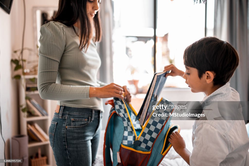 Young boy putting his notebook in the backpack, the mother is helping him