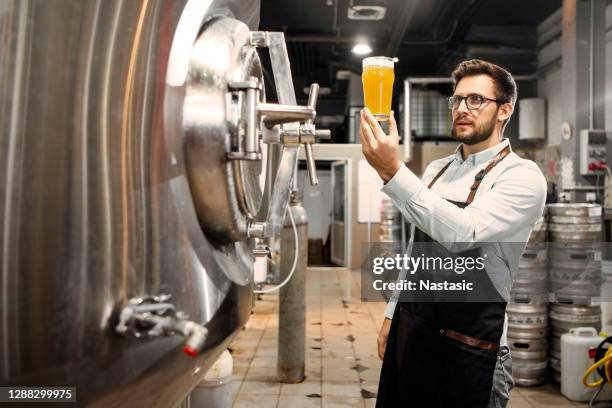 junger mann mit einem glas bier in brauerei-werk prüft qualität - brauerei stock-fotos und bilder