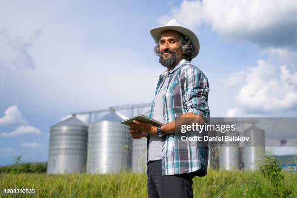 porträt eines bauern, der in der mitte eines weizenfeldes mit silos im hintergrund steht. landwirtschaftliche berufe. globales geschäft. investitionen in die landwirtschaft. - silo tank stock-fotos und bilder