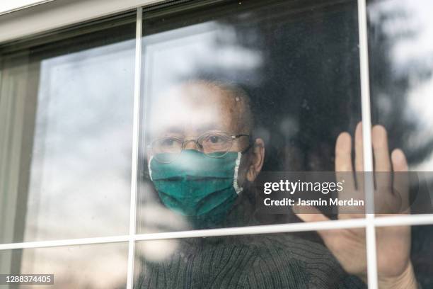 portrait of a senior woman wearing a face mask and looking through the window - looking through window covid stock pictures, royalty-free photos & images