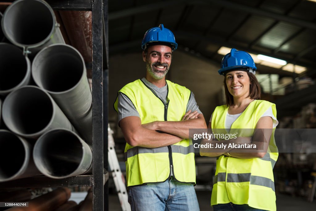 Man and woman pose looking at camera smiling near the tubes of construction material in a warehouse. Equal labor and rights. Teamwork image.