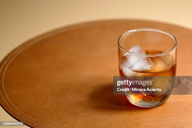 a glass of whisky and ice sit on a caramel color leather placemat. - whisky glass stock pictures, royalty-free photos & images