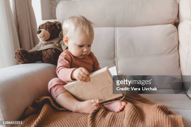 cute smiling little baby reading book sitting on sofa with teddy bear toy and bunny rabbit at home. - bébés garçons photos et images de collection
