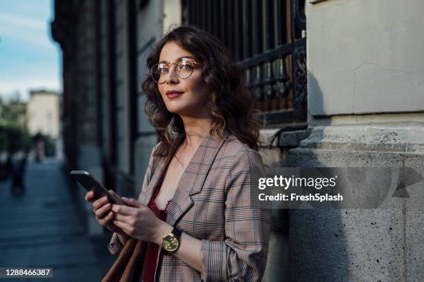 a beautiful woman standing in the street, holding her smartphone - elegancia imagens e fotografias de stock