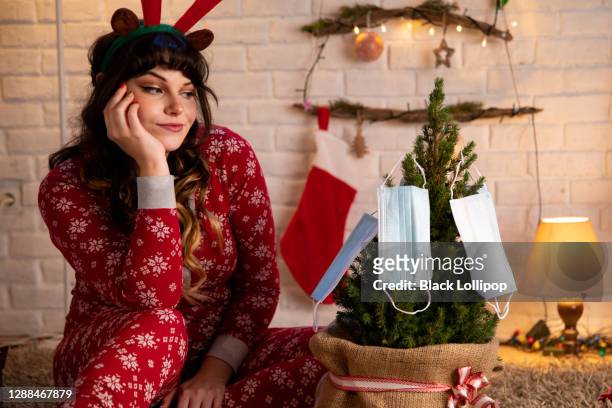 frau im winter pyjamas neben alternativen weihnachtsbaum mit schützenden gesichtsmasken. - funny surgical masks stock-fotos und bilder