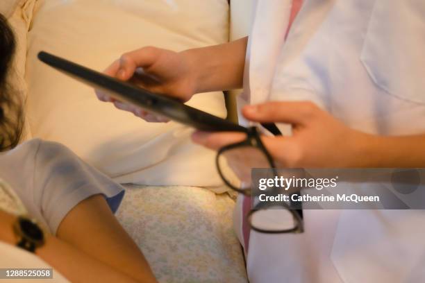 young mixed-race female doctor wearing pink medical scrubs & white lab coat at patient bedside holding eyeglasses & digital tablet - labbett stock-fotos und bilder