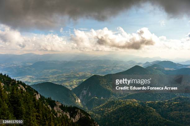 sunlight illuminating valley, forest and the rocky cliffs of piatra craiului mountains with a village and hills on the background, against a cloudy sky - transylvania stock pictures, royalty-free photos & images