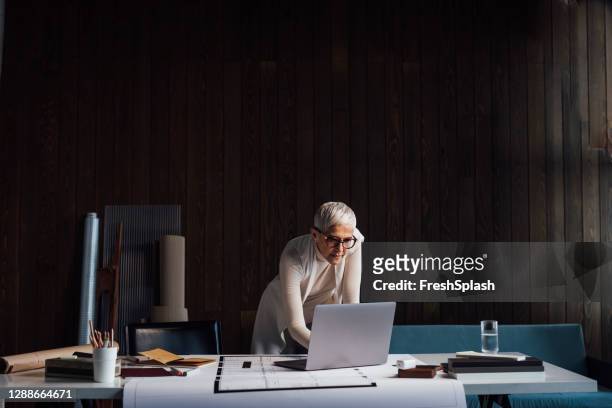 erfahrene ältere designerin, die an einem großen neuen projekt arbeitet und ihren laptop-pc in ihrem heimstudio verwendet (dunkler hintergrund) - eleganz stock-fotos und bilder