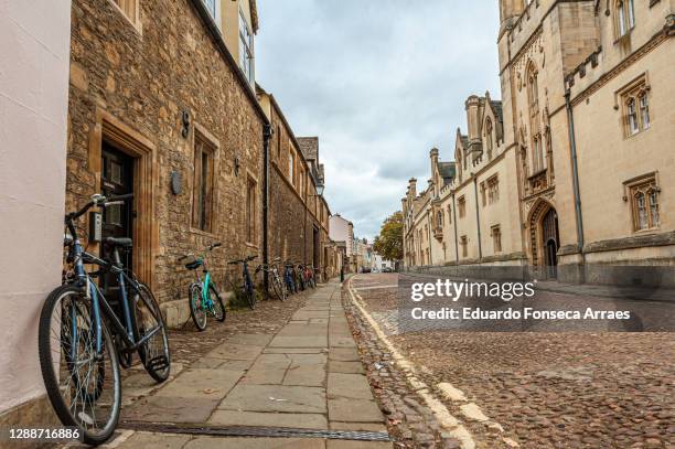 old cobblestone street lined by stone buildings and bicycles, against an overcast sky - oxford england stock pictures, royalty-free photos & images