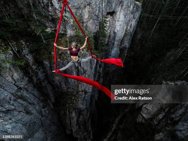 a female acrobatic does the splits on a red aerial silk that is suspended from a high-line spanning a tall rock gully face - acrobatic activity fotografías e imágenes de stock