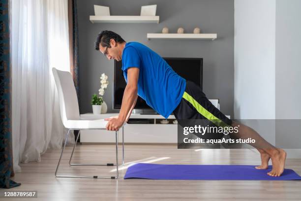 de jonge atletische mens doet duw omhoog oefening met stoel thuis in zijn ruime woonkamer met modern binnenland. gymnastiek thuis - chair exercise stockfoto's en -beelden