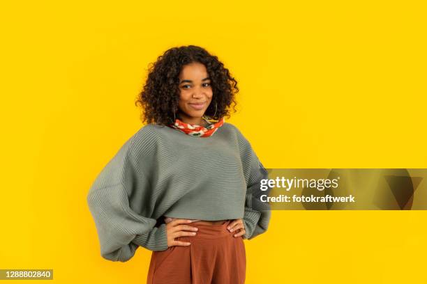 portrait of smiling young woman standing with hands on waist against yellow background - hand on hip stock pictures, royalty-free photos & images