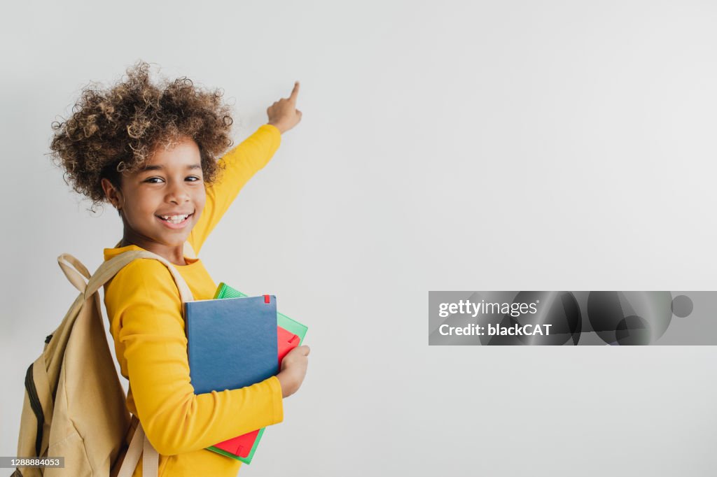 Garota fofinha afro-americana está segurando livros escolares um apontando em um espaço de cópia