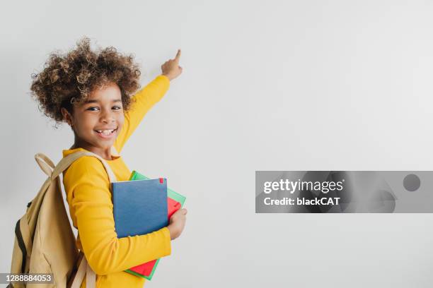 garota fofinha afro-americana está segurando livros escolares um apontando em um espaço de cópia - apontando sinal manual - fotografias e filmes do acervo