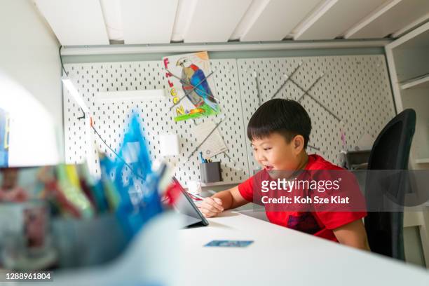 Young Asian boy sits at his desk under a bunk bed watching live stream on his digital tablet